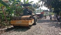 Alat berat Vibro Roller sedang memadatkan jalan berbatu di Desa Temmabarang, Kecamatan Penrang, Kabupaten Wajo, dalam program TMMD ke-124, dikelilingi oleh pohon pisang dan beberapa warga setempat.