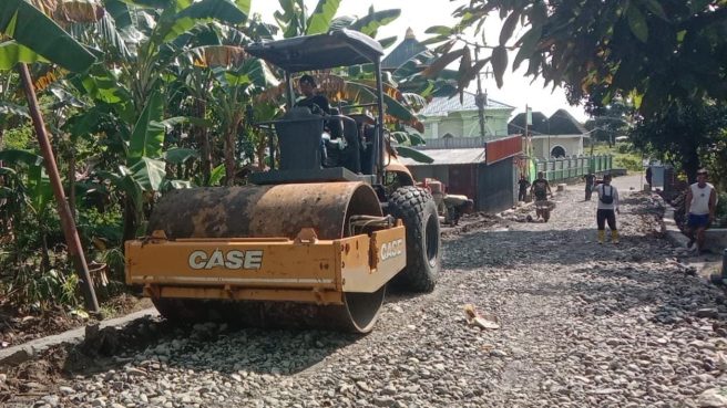 Alat berat Vibro Roller sedang memadatkan jalan berbatu di Desa Temmabarang, Kecamatan Penrang, Kabupaten Wajo, dalam program TMMD ke-124, dikelilingi oleh pohon pisang dan beberapa warga setempat.