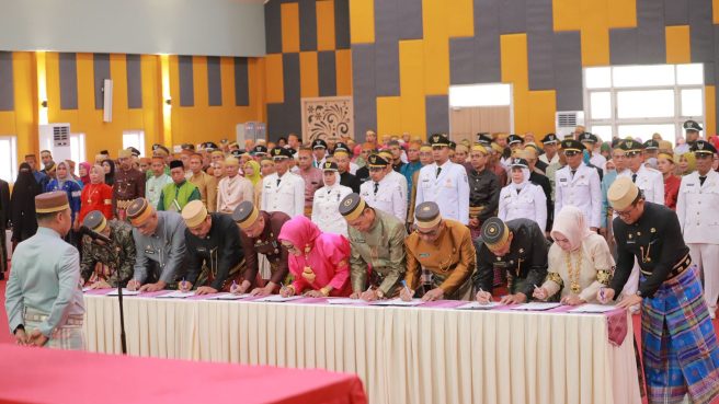 Sekelompok pejabat berpakaian adat Sulawesi Selatan dan seragam dinas sedang menandatangani dokumen secara massal di meja panjang dalam sebuah upacara formal yang dihadiri banyak orang.