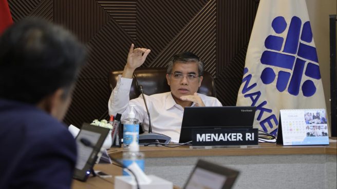 Menteri Ketenagakerjaan (Menaker) Yassierli duduk di ruang rapat Kemnaker mengenakan kemeja putih, memberikan penjelasan dengan gestur tangan, di depannya ada laptop dan papan nama 'MENAKER RI'.